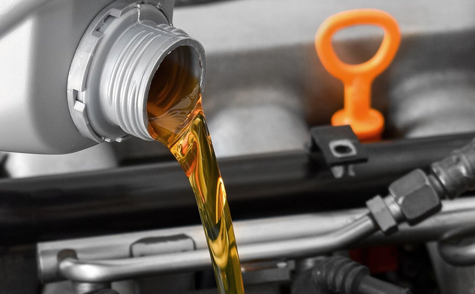 Engine oil being poured into a vehicle engine with an oil dipstick in the background.